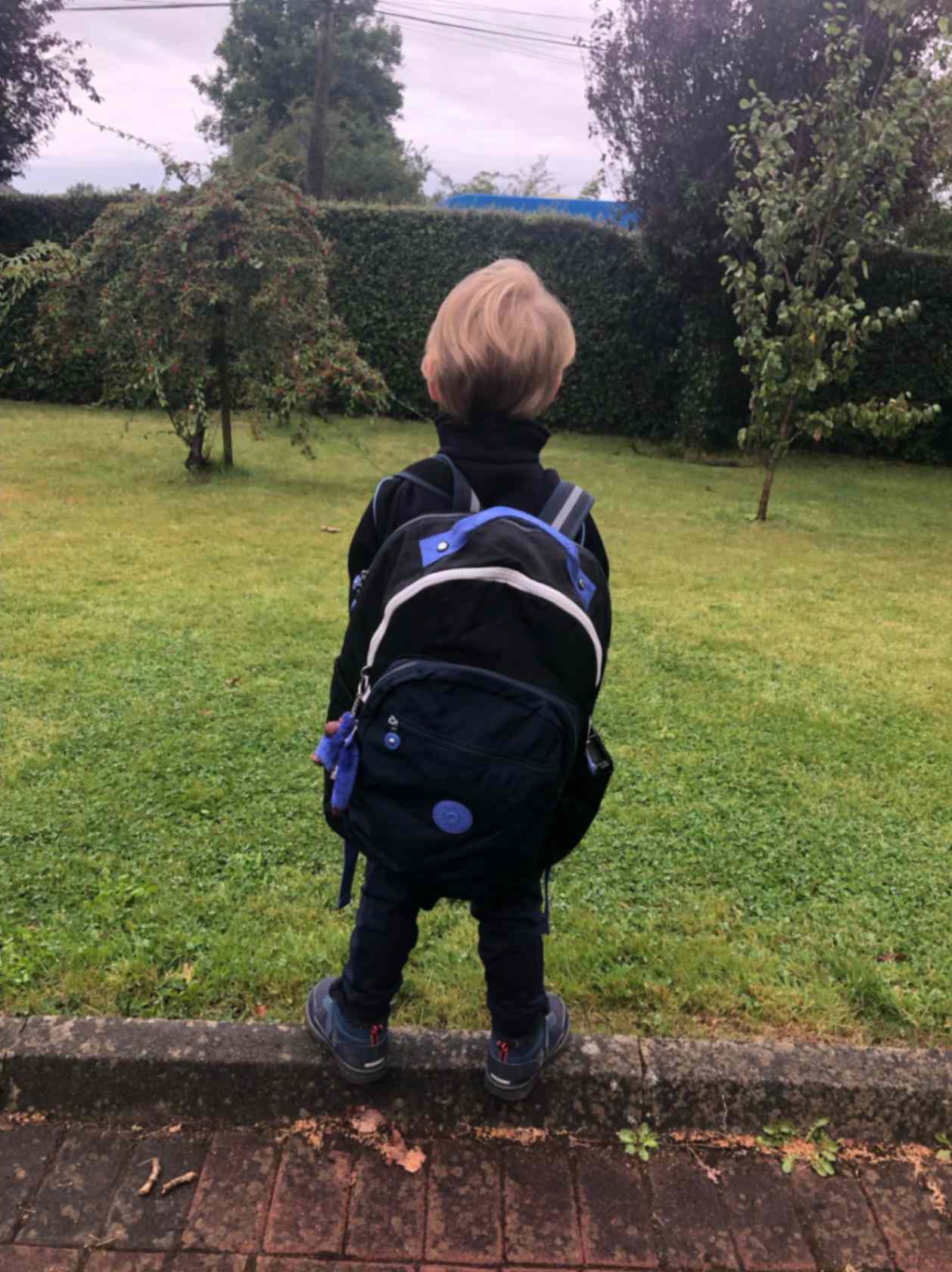 child-with-blonde-hair-stands-in-front-of-grass-wearing-a-large-black-and-blue-school-backpack