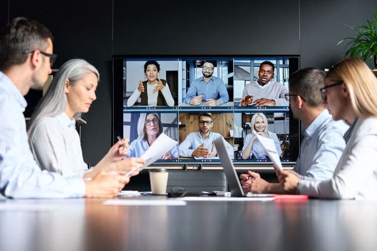 A group of people sitting at a conference table are engaged in a video call, as displayed on a large screen in front of them. The screen features multiple participants, including various individuals who are sharing ideas and discussing, some using hand gestures. The foreground shows a few people holding documents, while a coffee cup is placed on the table. The setting appears professional, with a modern aesthetic enhanced by a green plant in the corner.
