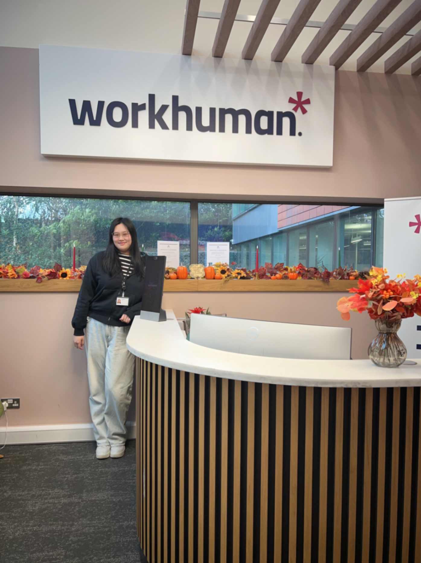 A woman stands next to a reception desk in front of a large sign that reads "workhuman." The desk has a curved surface with a wooden and black striped design. On the desk, there’s a computer screen and a small vase with flowers. In the background, there's a window with a view that includes decorative autumn-themed items, such as pumpkins. The setting appears welcoming and well-decorated.