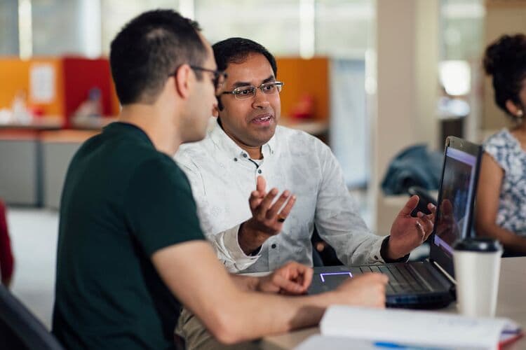 Two male coworkers are engaged in a discussion at a table around the topic of AI adoption, with one man wearing glasses and a white patterned shirt animatedly gesturing while speaking. The other man, wearing a green t-shirt, is listening attentively. In the background, a diverse group of people can be seen working at their own tables, with colorful office partitions present in the environment.