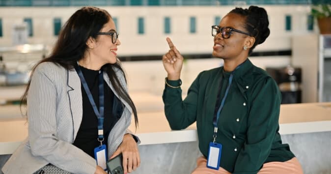 Two women engaged in a lively conversation while seated at a table. One woman, wearing a green blouse and glasses, is animatedly gesturing with her hand, while the other woman, dressed in a striped blazer, is smiling and listening attentively. Both women have identification badges hanging around their necks, and the background suggests a modern, professional environment.
