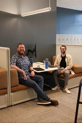 Two men are sitting in a modern, collaborative workspace. They are both smiling and appear to be engaged in conversation. One man, with a beard and wearing a blue patterned shirt, is sitting on the left, while the other, wearing glasses and a beige cardigan, is on the right. In front of them is a shared table that has a laptop and a blue water bottle. The backdrop features a wall with lockers. The overall atmosphere is casual and inviting.