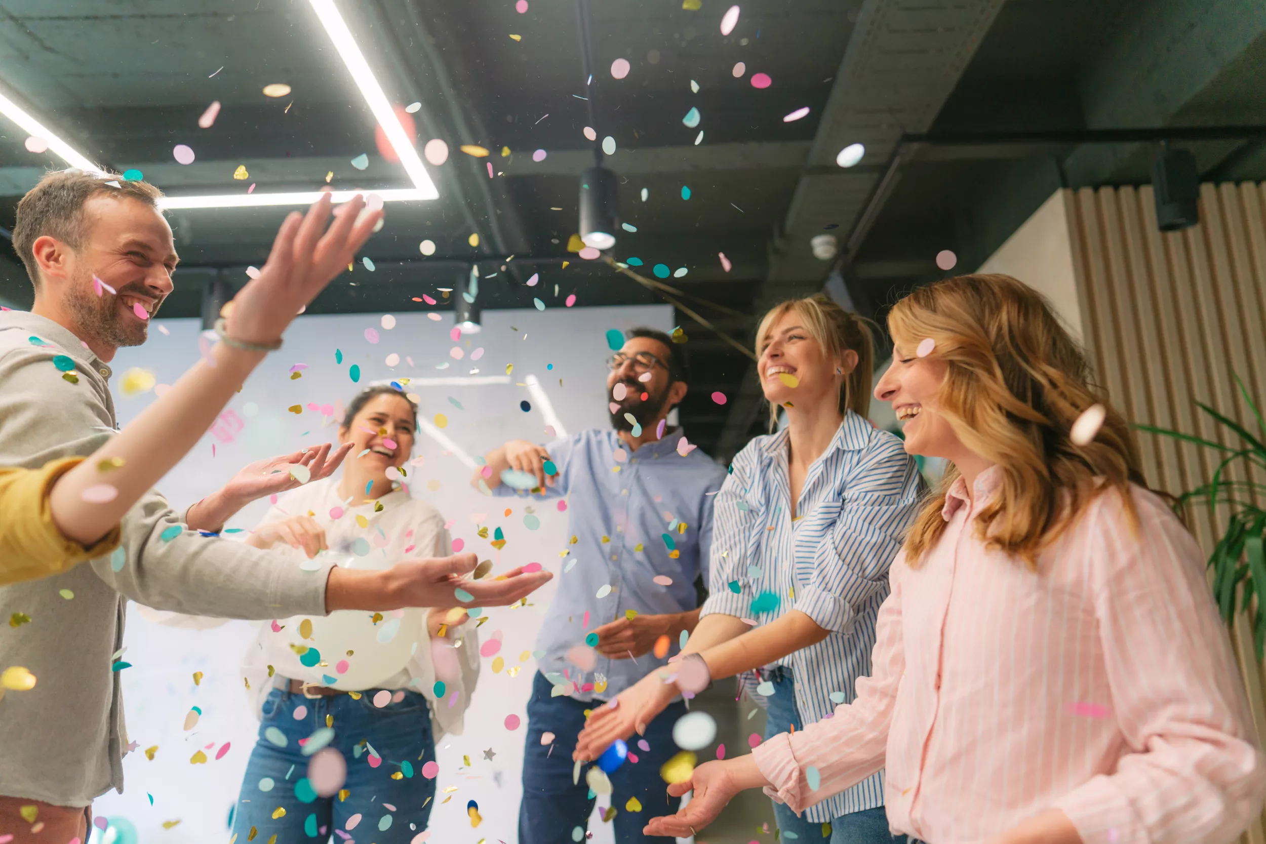 A group of five people joyfully celebrating while confetti falls around them. They are in a well-lit, modern space, with soft pastel colors visible in the confetti. The individuals are smiling, with arms outstretched as they engage in the celebration.