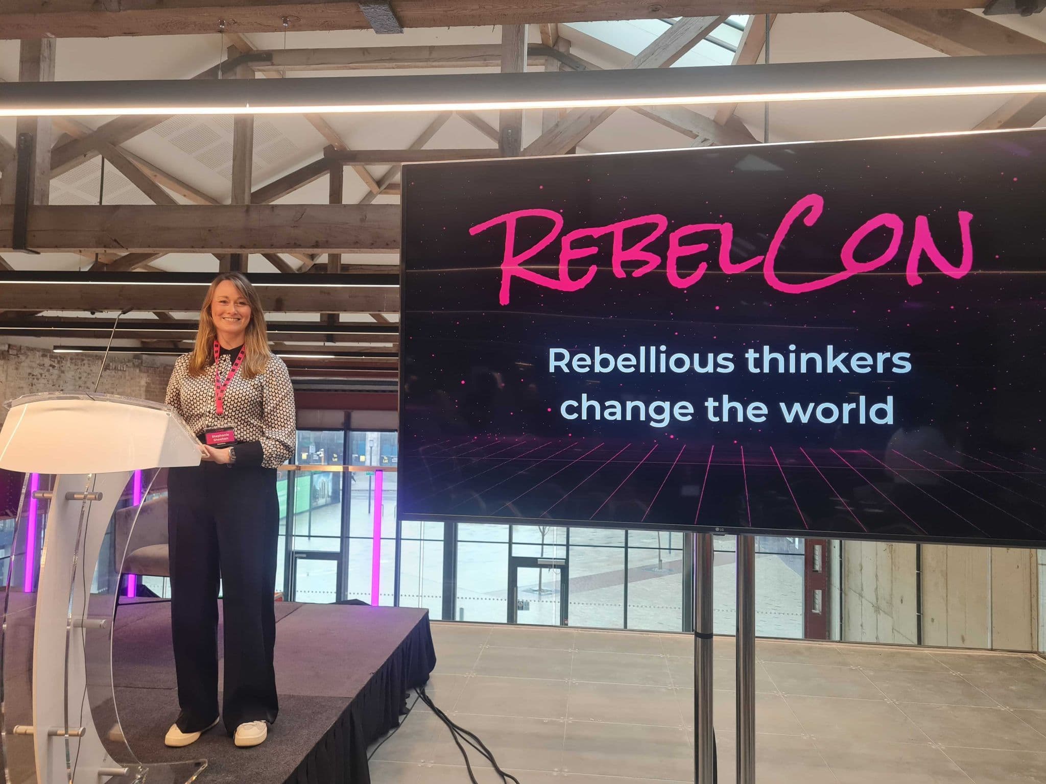 A speaker stands at a podium smiling while presenting at a conference titled "RebelCon." Behind her is a large screen displaying the event title "RebelCon" in bold, pink lettering and the tagline "Rebellious thinkers change the world." The setting features industrial-style architecture with visible wooden beams and large windows allowing light into the space.