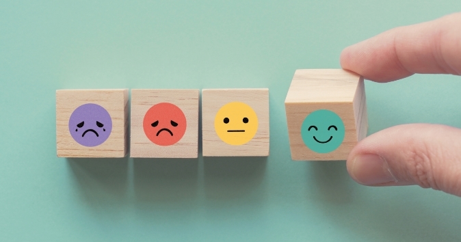 A hand is reaching for a wooden block decorated with a smiling face, positioned among three other blocks with various expressions: a sad purple face, a sad red face, and a neutral yellow face. The blocks are set against a soft green background.