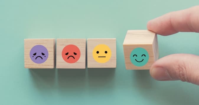 A hand is reaching for a wooden block decorated with a smiling face, positioned among three other blocks with various expressions: a sad purple face, a sad red face, and a neutral yellow face. The blocks are set against a soft green background.