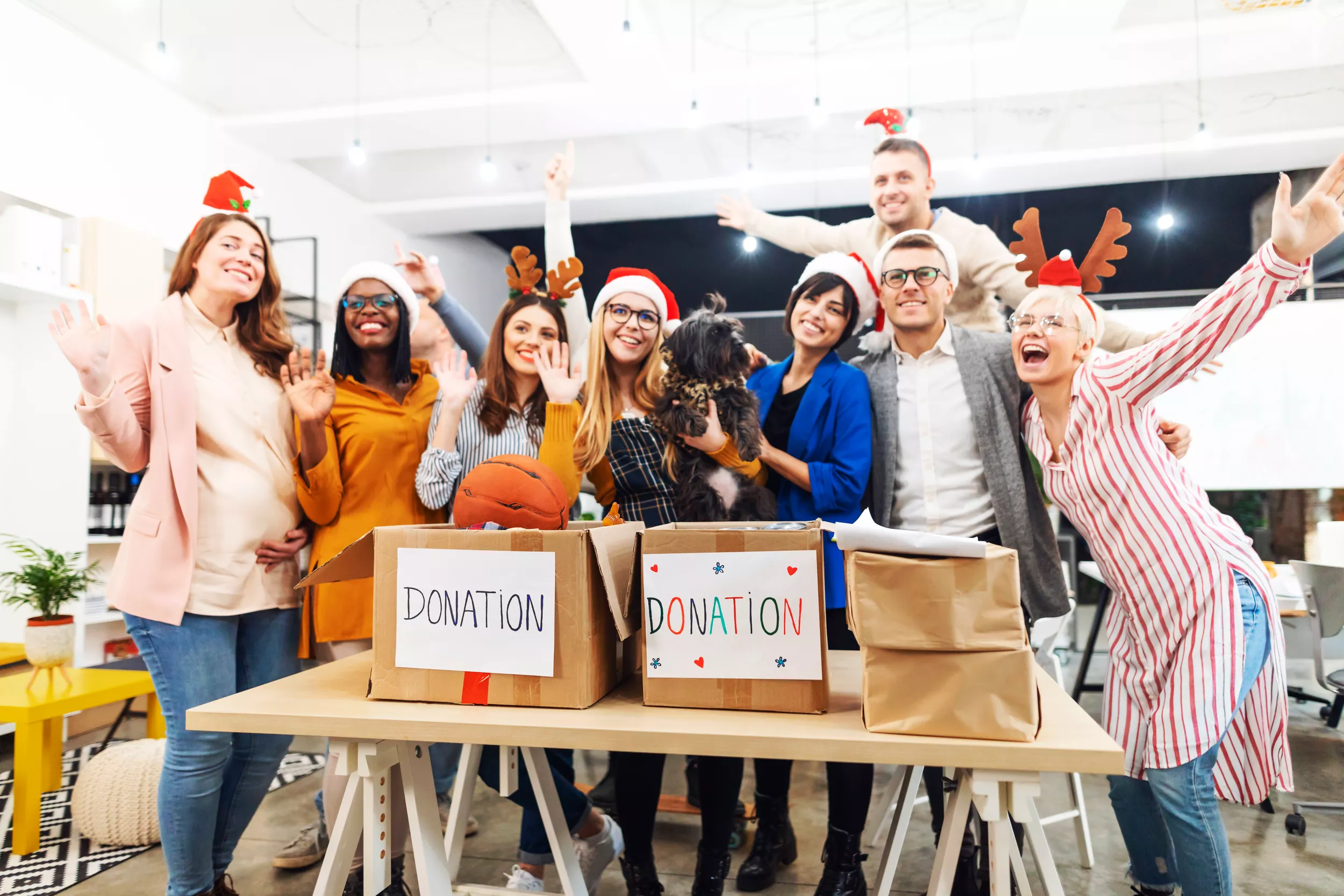 A cheerful group of eight individuals, wearing festive hats, stand around a table filled with donation boxes, each marked with the word "DONATION." They are smiling and waving, showcasing a joyful atmosphere in a bright, modern workspace. One person is holding a small dog, adding to the lively scene.