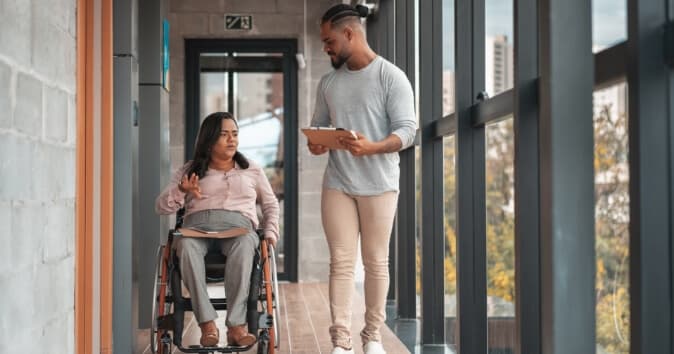 Woman in a wheelchair going down a hall with a man walking alongside of her.