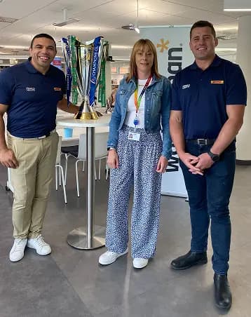Three individuals are smiling together in an indoor setting, standing next to a trophy on a pedestal. Two men, dressed in matching navy polo shirts, are positioned on either side of a woman wearing a light blue jacket and patterned pants. A banner in the background features the Workhuman logo, and colorful ribbons are visible nearby. The atmosphere appears celebratory and engaging.