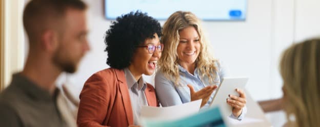 A group of four colleagues is engaged in a discussion in a modern office setting. Two women in the foreground, one with curly hair wearing glasses and an orange blazer, excitedly show each other documents and a tablet. The other two colleagues, a man in a light-colored shirt and a woman in a blue blouse, observe and listen. In the background, a screen displays a financial graph, contributing to the professional atmosphere.