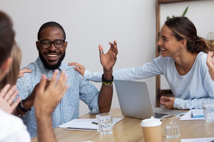 A group of colleagues celebrates together in a meeting room. One man, wearing glasses and a patterned shirt, sits at a table with a laptop and water glasses. He appears joyful as a woman beside him gestures expressively, and another colleague reaches out to him, creating a lively atmosphere of teamwork and support.