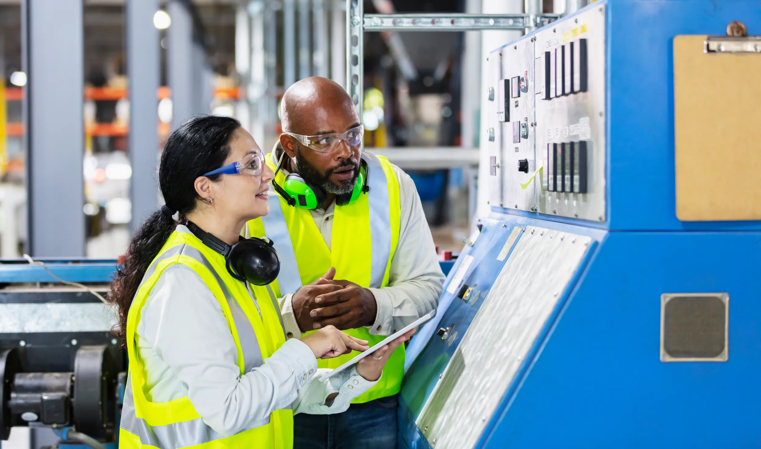 A woman and a man are collaborating in a factory setting, both wearing bright yellow safety vests and protective eyewear. The woman holds a tablet, pointing at a control panel on a large blue machine, while the man, who has a short beard and wears headphones, looks on attentively. The background features a large industrial space with racks and machinery.