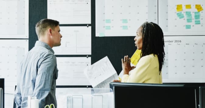 Two people in conversation near a large wall of printouts.