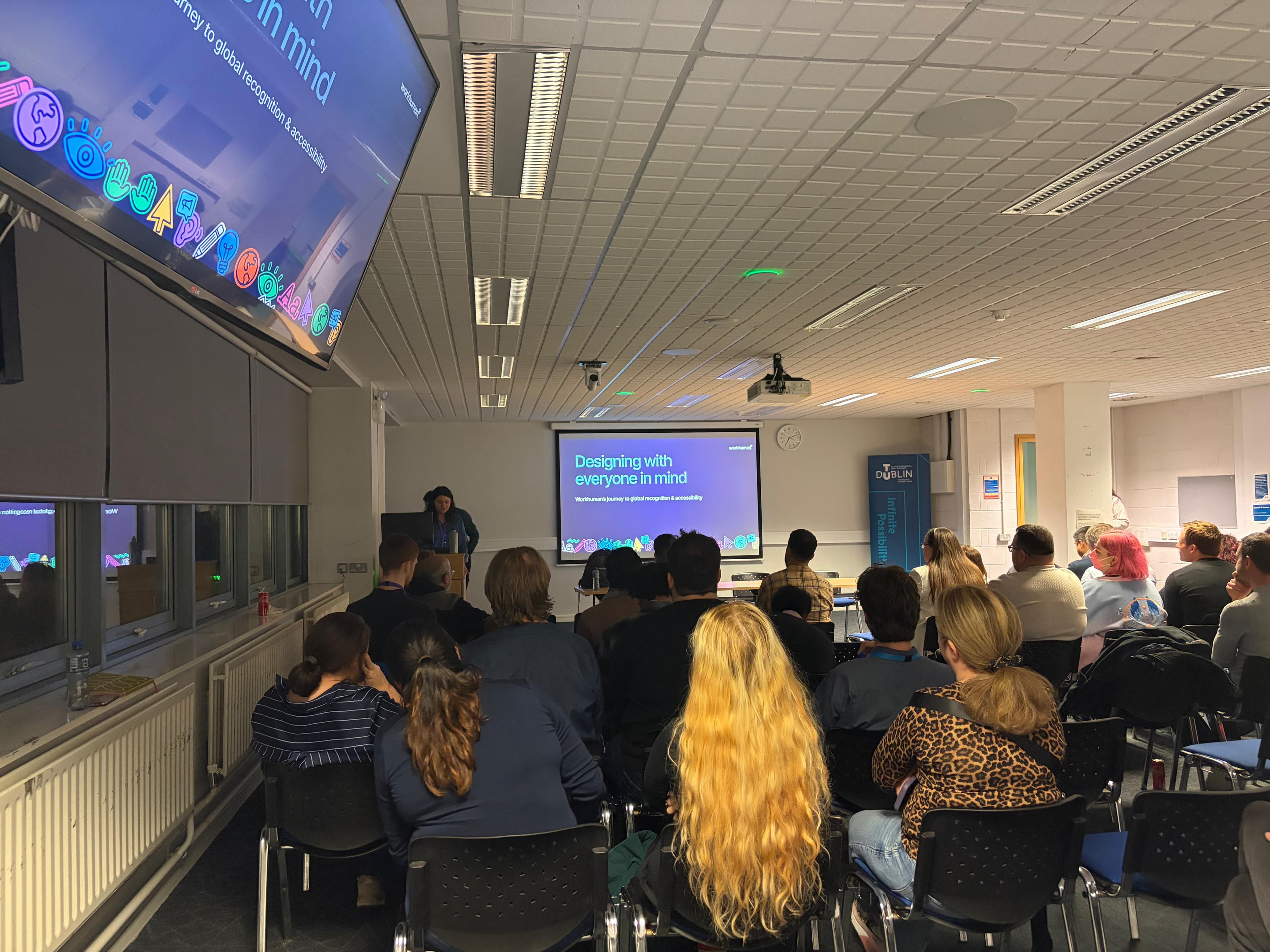 A speaker stands at a podium in front of an audience during a presentation titled "Designing with Everyone in Mind," highlighting Workhuman's journey towards global recognition and accessibility. The slide displayed on a large screen features colorful icons representing inclusivity and accessibility. Attendees are seated, focused on the presentation, with some visible reactions to the content.