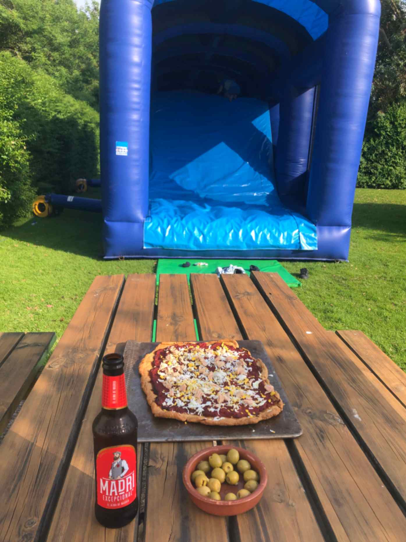 a-wooden-table-with-a-pizza-a bottle of beer-and-a-bowl-of-olives-with-a-bouncy-castle-in-the-background