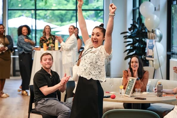 A jubilant woman raises her arms in celebration at a gathering, surrounded by several attendees who are smiling and clapping. The atmosphere appears joyful, with drinks on tables and a bright, airy setting featuring large windows. The backdrop includes potted plants and a festive arrangement for the event.