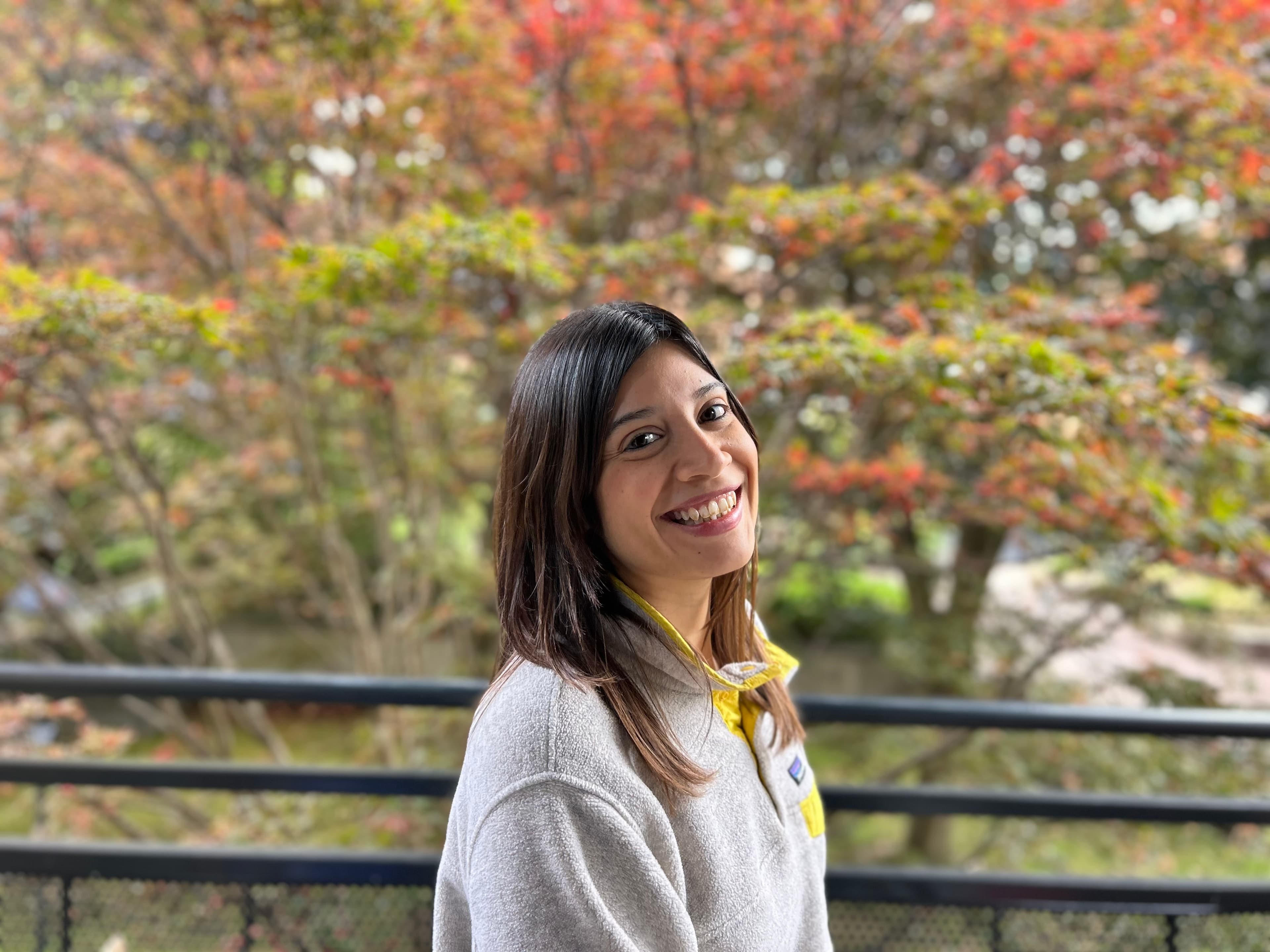 A smiling woman with long hair is standing outside, wearing a light-colored sweater with a yellow collar. She is looking towards the camera, with a bright smile. In the background, there are colorful trees, displaying shades of green and red foliage, creating a serene outdoor setting.