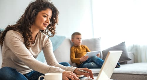 woman sitting on a couch working on a laptop with a child nearby