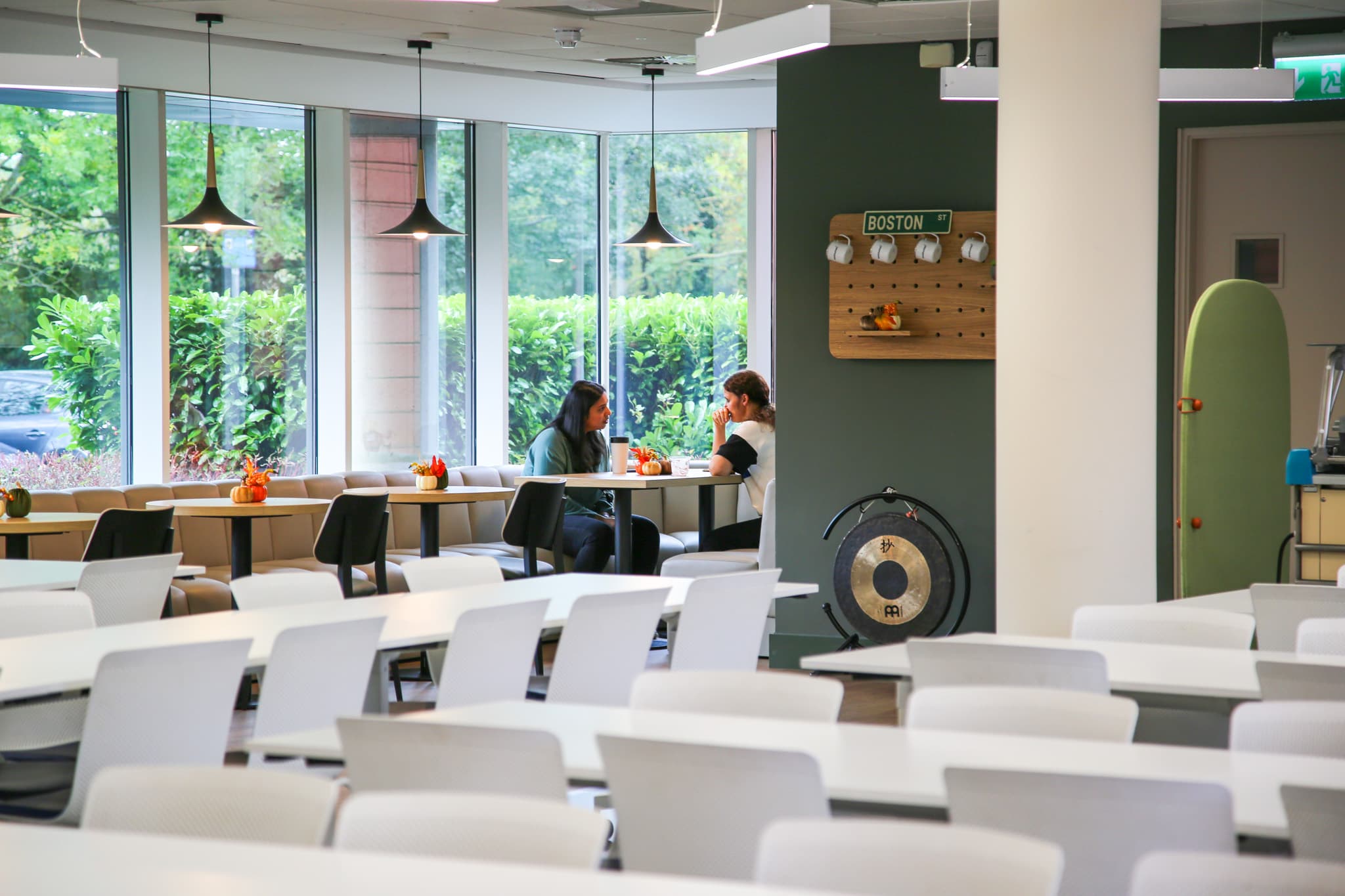 Two women are seated at a table in a bright, modern café space with large windows. They are engaged in conversation over drinks, while small decorative pumpkins adorn their table. The background features various empty tables and chairs, a wall-mounted display with mugs, and a gong. Large greenery is visible outside the windows, indicating a well-lit, inviting atmosphere.
