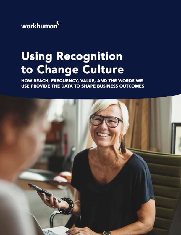 A smiling woman with glasses is sitting at a table, holding a smartphone. She is engaged in conversation with another person partially visible on the left. In the background, there are soft furnishings and a modern office setting. The image prominently features text that reads, "Using Recognition to Change Culture: HOW REACH, FREQUENCY, VALUE, AND THE WORDS WE USE PROVIDE THE DATA TO SHAPE BUSINESS OUTCOMES," along with the Workhuman logo.