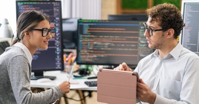 A woman and a man wearing glasses are engaged in a discussion at a workspace filled with multiple computer monitors displaying lines of code. The woman is holding a pen and looking attentively at the man, who is holding a tablet and explaining something. The environment reflects a collaborative tech setting with a blend of personal touches and a focus on technology.