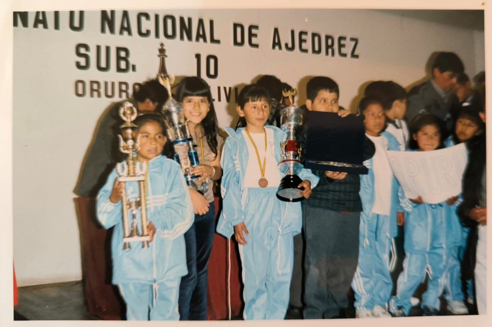 A group of children gathered at a chess competition event, holding trophies and medals. The front row features several children dressed in matching light blue tracksuits, three of whom are holding large trophies, one showing a medal around their neck. The backdrop includes a banner that reads "NAIU NACIONAL DE AJEDREZ SUB. 10 ORUR," indicating the national chess tournament for under-10 participants in Oruro, Bolivia. Other children stand behind them, some holding a sign. The scene captures the excitement and achievement of the event.