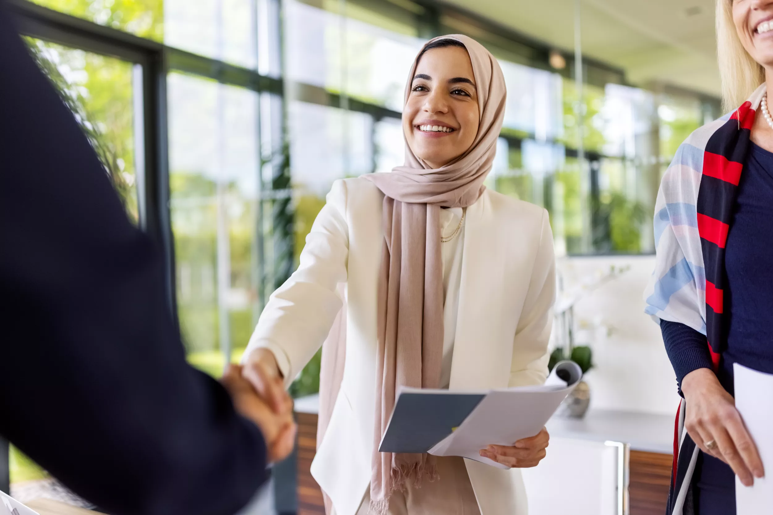 A smiling woman wearing a beige hijab stands in a bright indoor setting, extending her hand to shake hands with another person. She holds documents in her left hand and is dressed in a white jacket. To her right, another woman is partially visible, also dressed professionally. Large windows behind them provide a view of greenery outside.