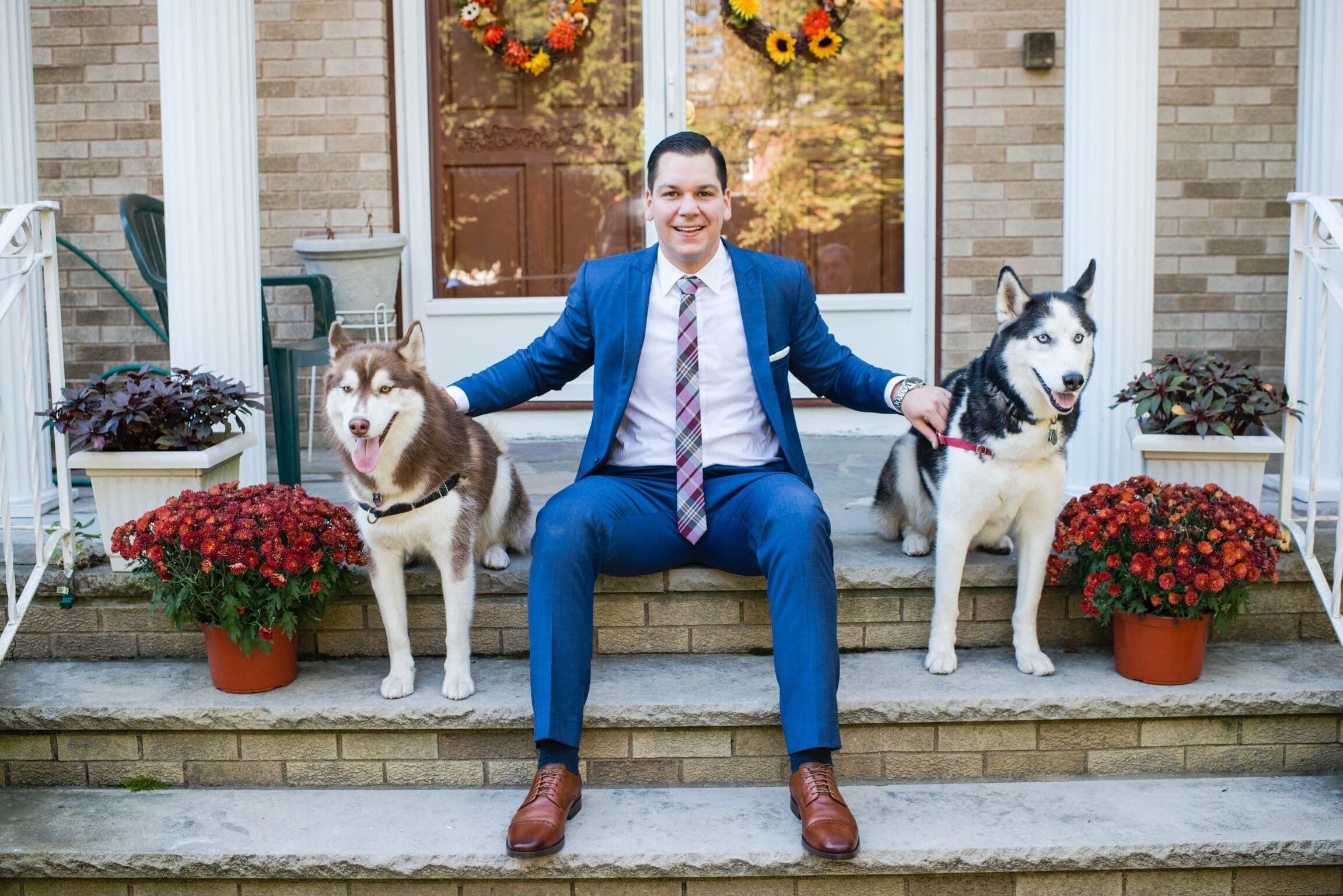 A man in a blue suit sits on the steps of a house, smiling and posing with two Siberian Huskies—one brown and white, and the other black and white. They are flanked by flower pots with orange flowers. The backdrop features a wooden door adorned with a seasonal wreath.
