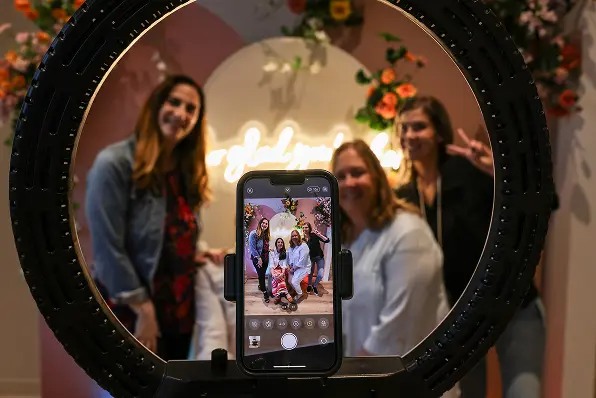 A group of three women poses for a photo in front of a floral backdrop while another woman captures the moment with a smartphone. The phone, positioned within a ring light, displays the image of the group. The backdrop features a circular design with soft colors and decorative flowers, creating a festive atmosphere.