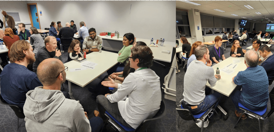 A group of people engaged in conversation at tables in a meeting or conference setting. Two separate groups are visible; one on the left side appears to have a mix of men and women, while the group on the right side is primarily men. The atmosphere is interactive, with attendees discussing whatever is on the printed sheets in front of them. Beverages are present on the tables, and the setting is well-lit with modern furnishings.