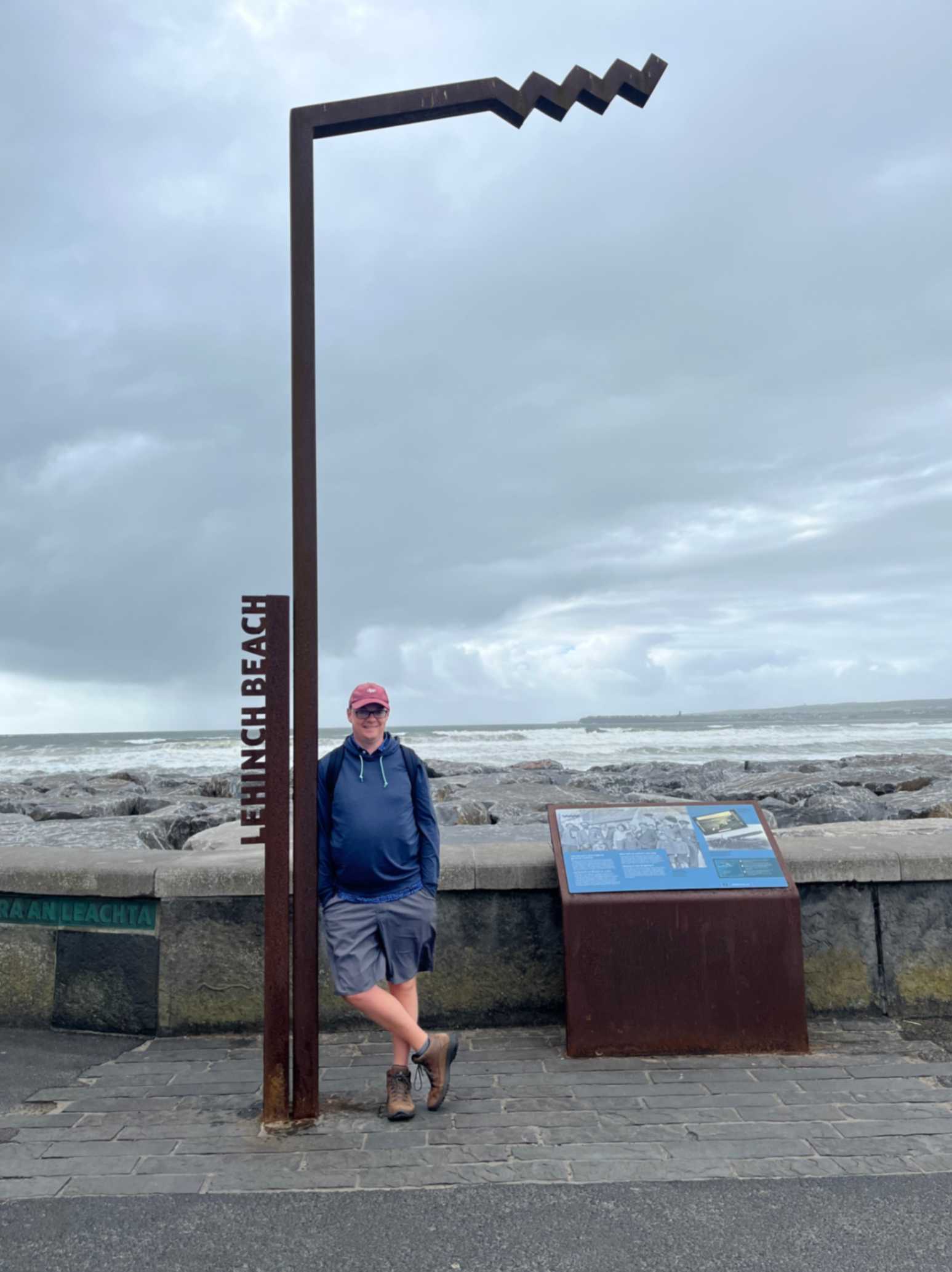 A person stands next to a large, angular metal sign that reads "Lahinch Beach." They are wearing a blue hoodie and shorts, with a backpack slung over one shoulder. Behind them is a rocky beach with waves crashing against the shore under a cloudy sky. To the side, there is an informational display featuring text and images about the area.