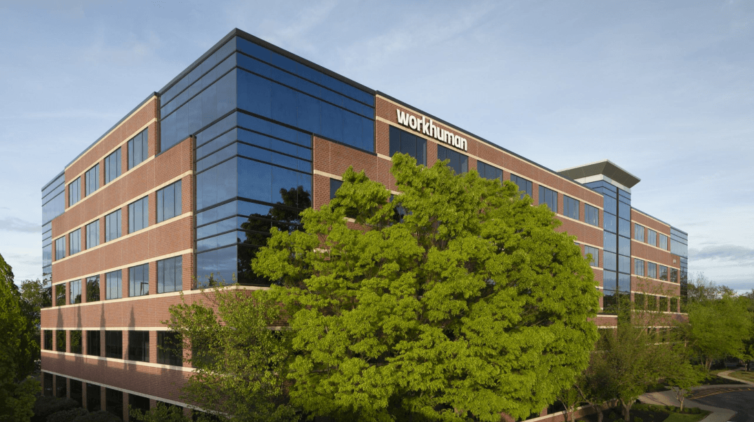 A modern office building with large glass windows and a brick exterior, prominently featuring the Workhuman logo at the top. Surrounding the building are lush green trees and landscaping, indicative of a well-maintained environment. The image is taken from an angle that showcases the corner of the building against a clear blue sky.