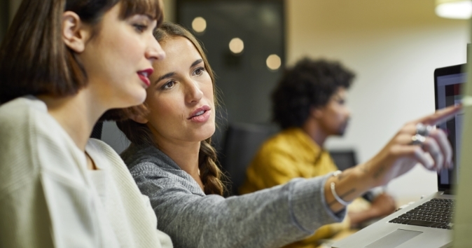 Two women are engaged in discussion, with one pointing towards a computer screen while the other listens attentively. The background features a man, slightly blurred, working at another computer station. The setting appears to be a collaborative workspace.