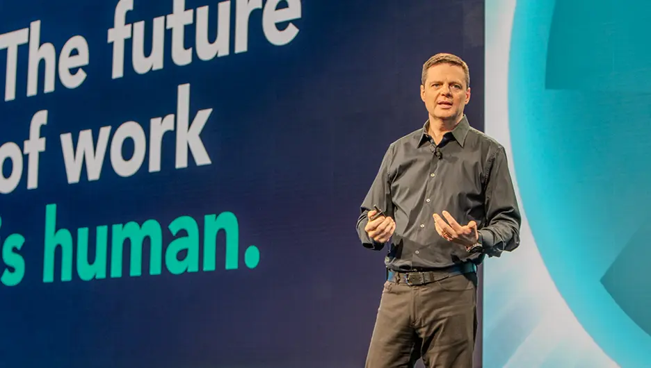 A speaker in a dark shirt stands in front of a large screen displaying the text, "The future of work is human." The background features a gradient design, and the speaker appears to be engaged in a presentation or discussion.