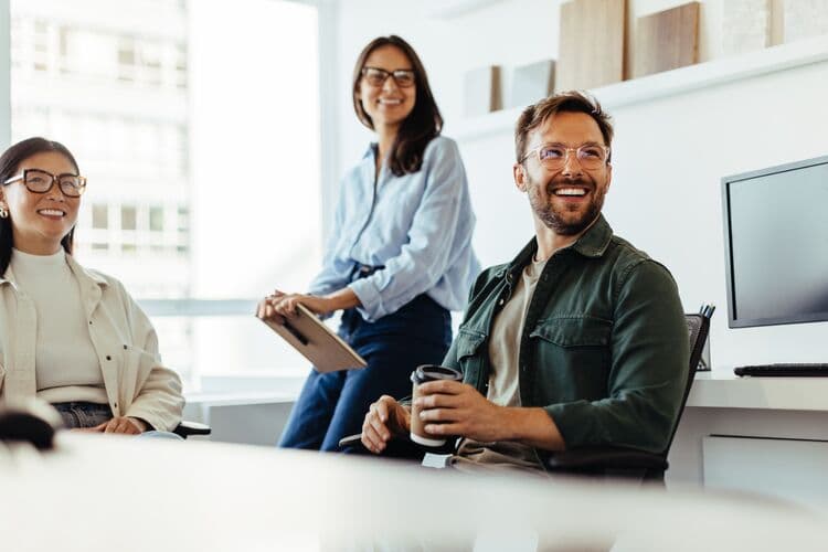 A group of three HR professionals are seated in a bright, modern office space. Two women, one wearing a white blouse and the other in a light blue shirt, are smiling. The man in the foreground, wearing glasses and a green jacket, holds a coffee cup and is also smiling. The atmosphere conveys a sense of teamwork and collaboration. A computer and various materials are visible in the background, contributing to the workspace setting. They are being celebrated on HR Professionals day.