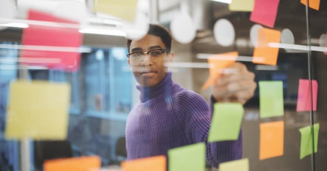 A young man wearing a purple sweater is standing in front of a glass wall covered with colorful sticky notes in various shapes and sizes that outline his talent management system.