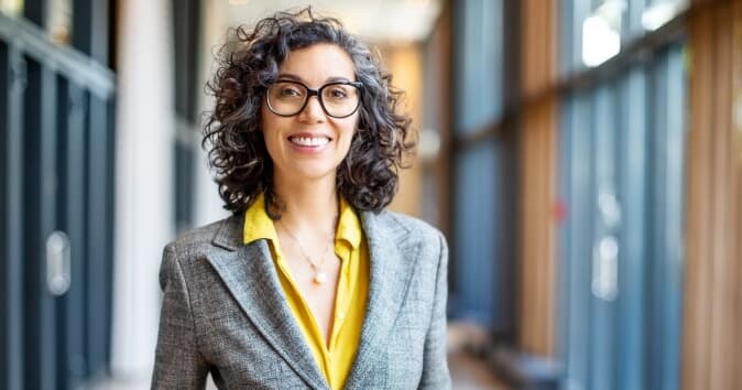 A smiling woman with curly hair is standing in a corridor, wearing a grey blazer over a yellow blouse. She has glasses and exudes a friendly demeanor, with wooden paneling and large glass windows visible in the background.