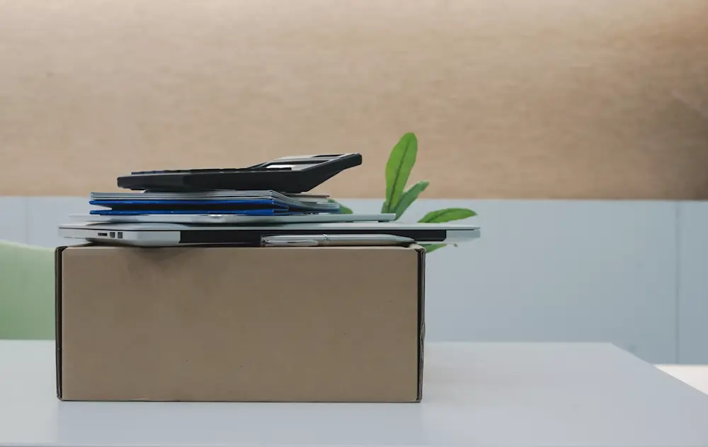 A stack of items resting on a cardboard box on a table, including a laptop, a notebook, a calculator, and a few other papers or documents. A green plant is visible next to the box, adding a touch of greenery to the workspace. The background features a neutral, light-colored wall.