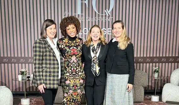 Four women are posing together and smiling in front of a stylish backdrop featuring the text "FQ" and "The Quotient Lounge." Each woman is dressed in different outfits, showcasing a blend of professional and casual styles. The setting appears to be a modern lounge area, with soft seating and decorative elements present in the background.