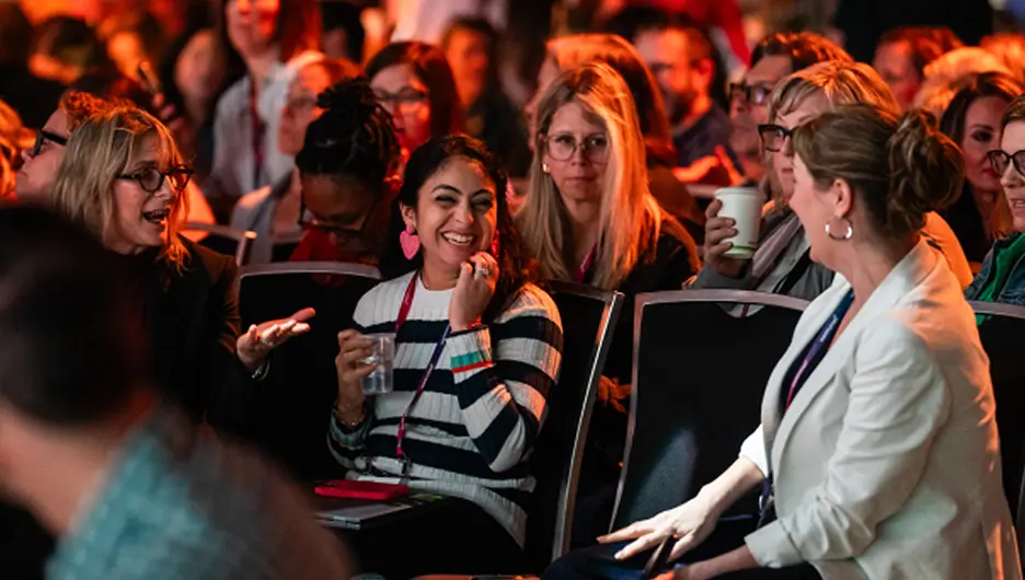 A group of diverse attendees at a conference are engaged in conversation, reflecting a lively atmosphere. In the foreground, a woman wearing a striped shirt is smiling and animatedly speaking with a companion, while several others around them are attentive and smiling, indicating camaraderie and excitement. The background is filled with people, some holding drinks, all contributing to the energetic environment of the event.