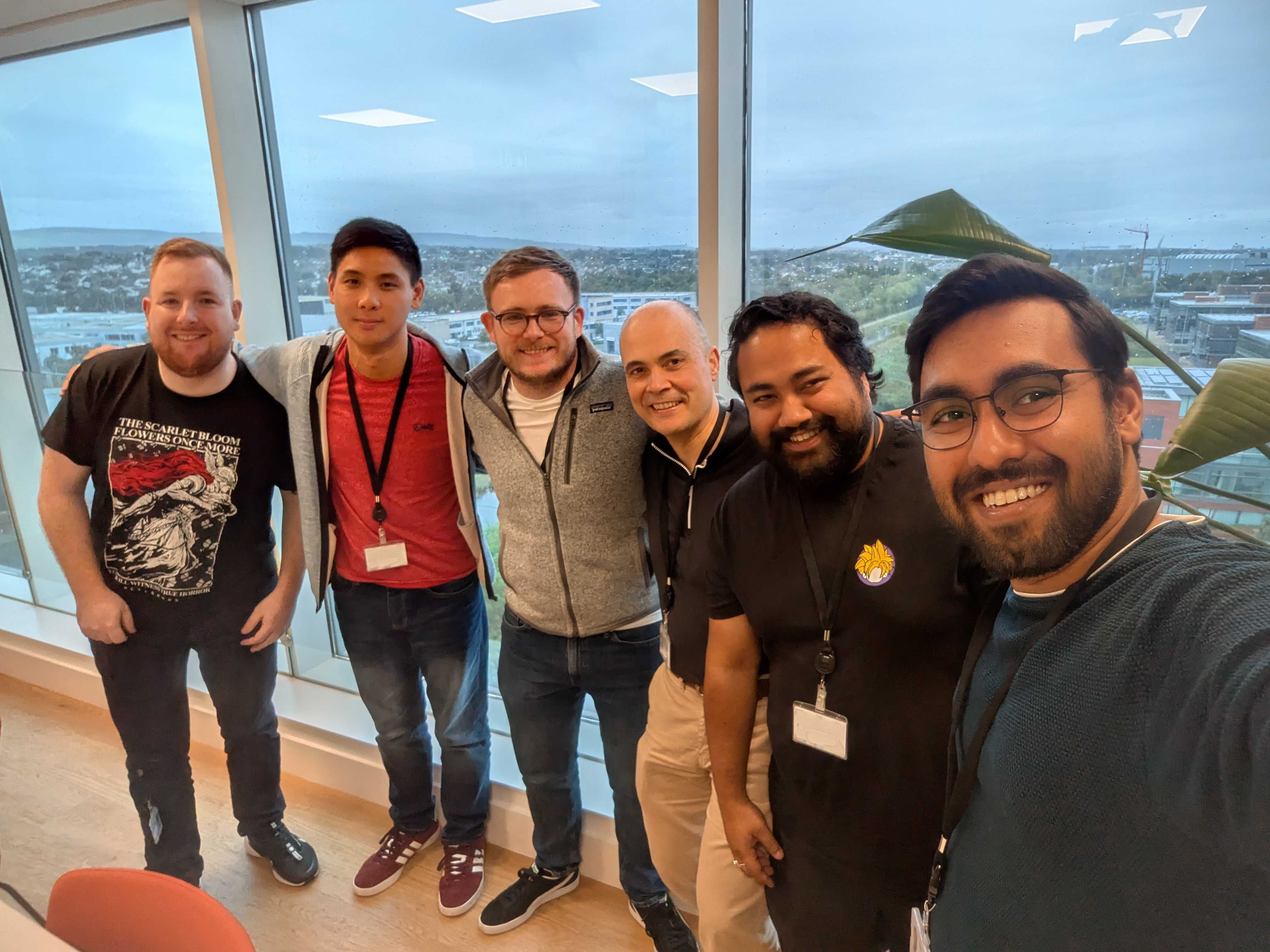 A group of five individuals is posing for a selfie indoors, facing the camera with smiles. They are standing in front of a large window that shows a cloudy landscape outside. The person on the far left is wearing a black t-shirt with an artistic design, while the others wear casual clothing, including a red shirt and a grey zip-up jacket. One person holds a name tag around their neck, and there are green plants in the background. The atmosphere appears friendly and casual, indicating a group gathering or event.