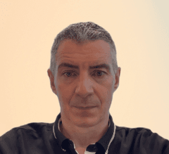 A man is looking directly at the camera, wearing a dark shirt. The background is a plain, light color, creating a neutral backdrop.