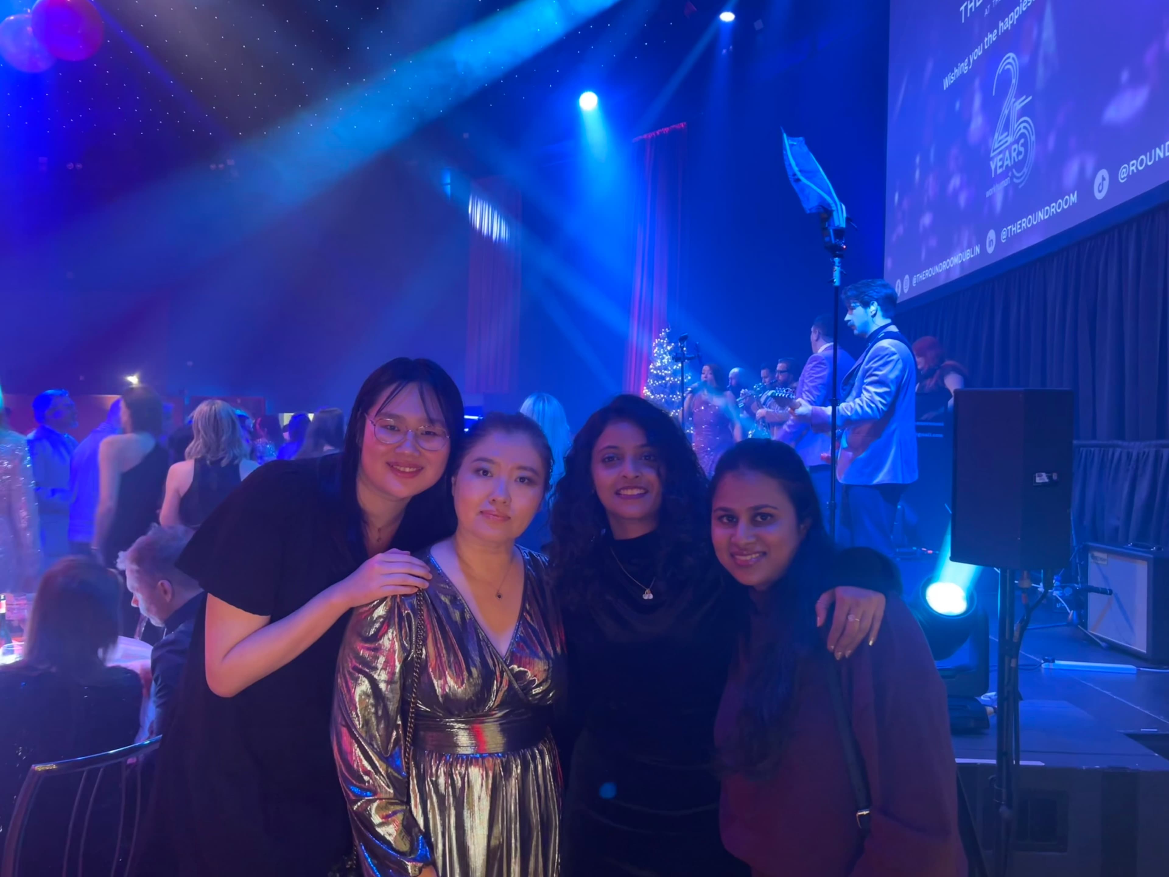 Four women pose together at an indoor event with blue lighting, surrounded by other attendees; a stage and screen are visible in the background.
