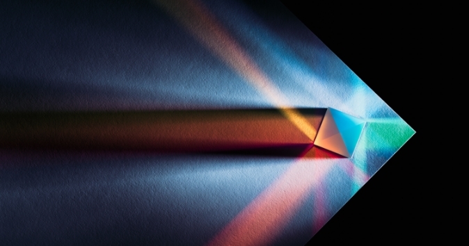 A prism casting colorful beams of light onto a textured surface. The light creates dynamic patterns with shades of blue, green, and orange as they spread out in various directions from the prism. The background is dark, enhancing the brightness and vibrance of the colors.