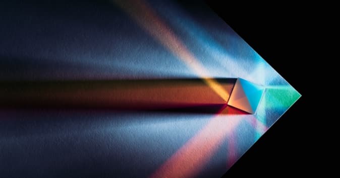 A prism casting colorful beams of light onto a textured surface. The light creates dynamic patterns with shades of blue, green, and orange as they spread out in various directions from the prism. The background is dark, enhancing the brightness and vibrance of the colors.