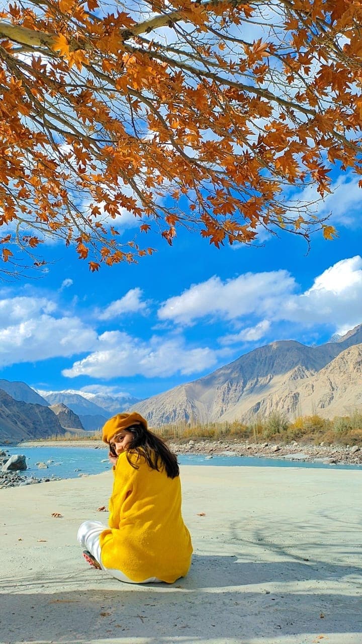 A person wearing a bright yellow sweater and hat sits on a sandy surface, looking back over their shoulder while smiling. They are surrounded by vibrant autumn foliage and a serene landscape featuring mountains and a clear blue sky. There is a river in the background, enhancing the picturesque nature of the scene.