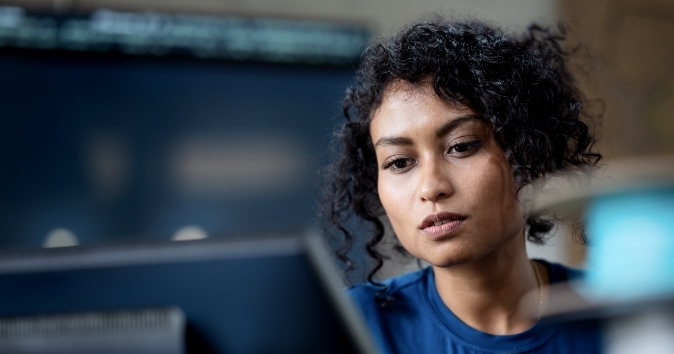 A woman with curly hair is focused on a computer screen, appearing to concentrate intently, while sitting in a workspace with a blurred background.