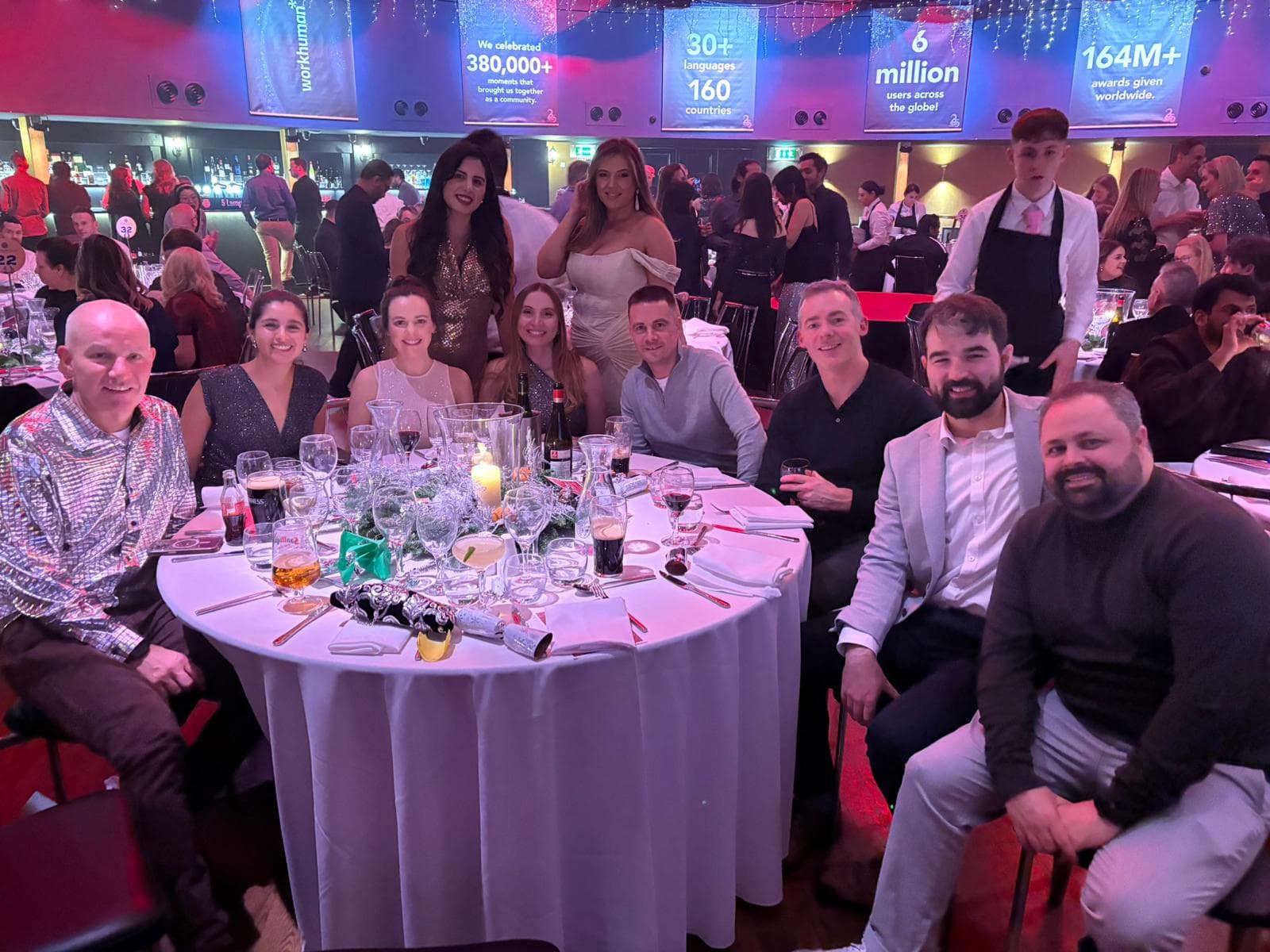 A group of people gathered around a table at a festive event, smiling and enjoying each other's company. The table is set with glasses, drinks, and decorative items. In the background, there are decorations and a waiter serving guests. Prominent text appears above them, highlighting achievements such as "We celebrated 380,000+ moments that brought us together as a community," along with figures indicating six million users and 164 million awards given globally.