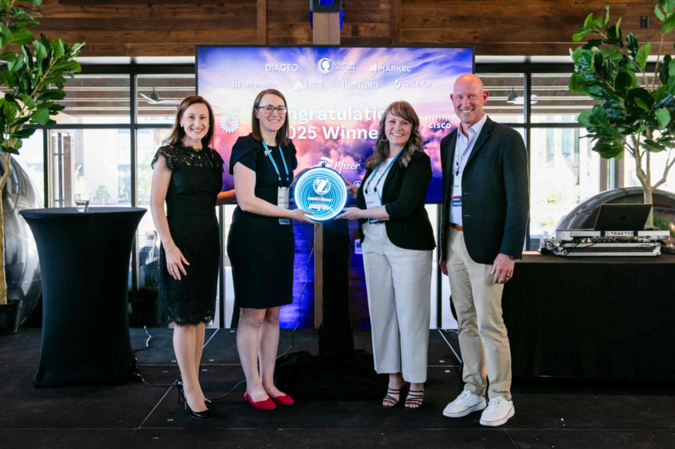 Four people stand on stage, smiling and holding a glowing award. A banner behind them reads "Congratulations 2025 Winners." The mood is celebratory.