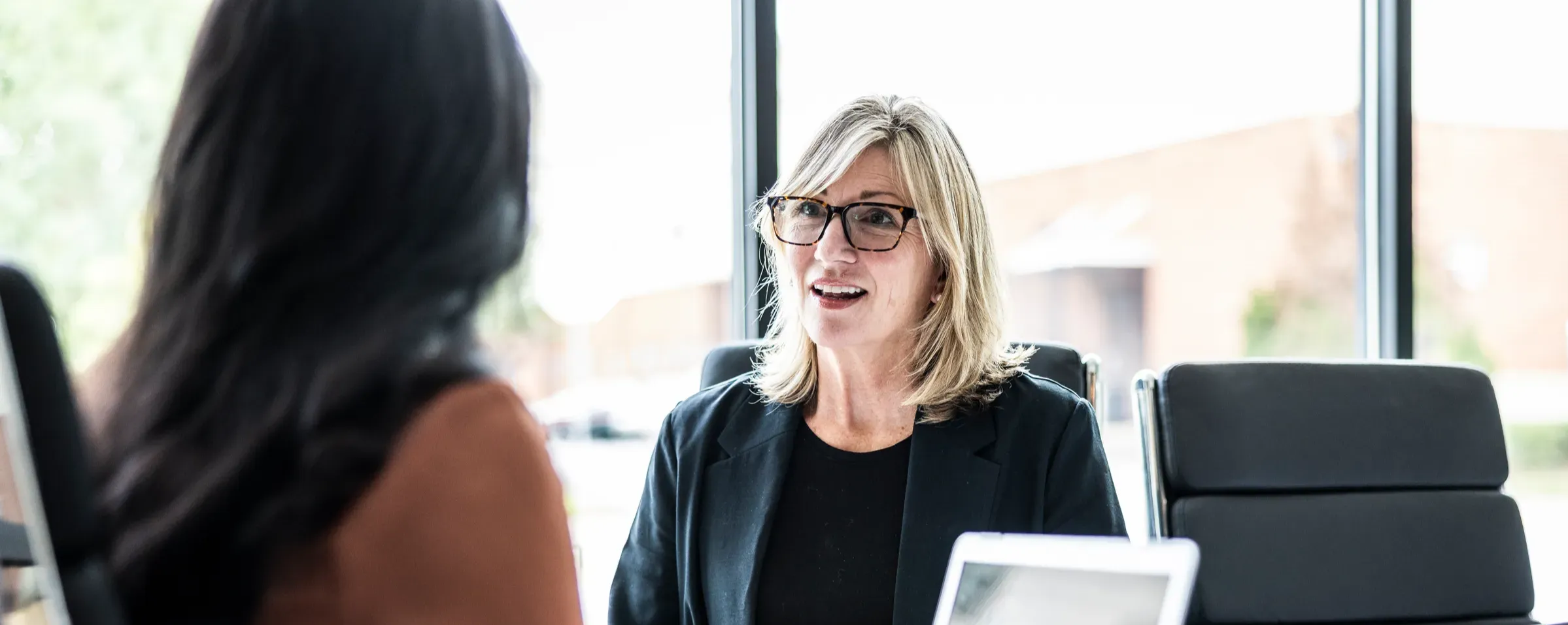 A woman with shoulder-length blonde hair, wearing glasses and a black blazer, is engaged in conversation with another person who is not fully visible. The setting appears to be a bright, modern office with large windows, behind which an industrial building is faintly seen. A laptop is positioned on the table between them.
